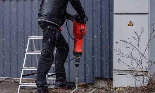 Elektricien slaat met een breekhamer een aardpen in de grond naast een kast